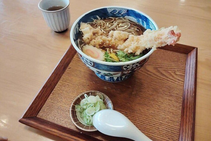 En skål med ramen, med en bit friterad fisk ovanpå, en liten skål med grönsaker och en vit sked på ett träbricka. 2