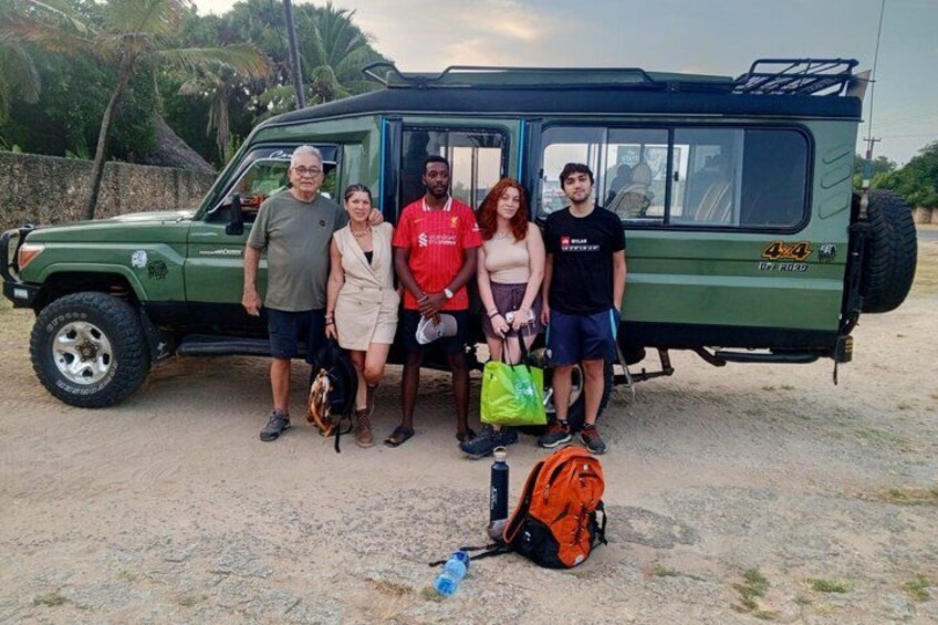 A group of people standing in front of a green vehicle with a luggage bag on the ground. 2