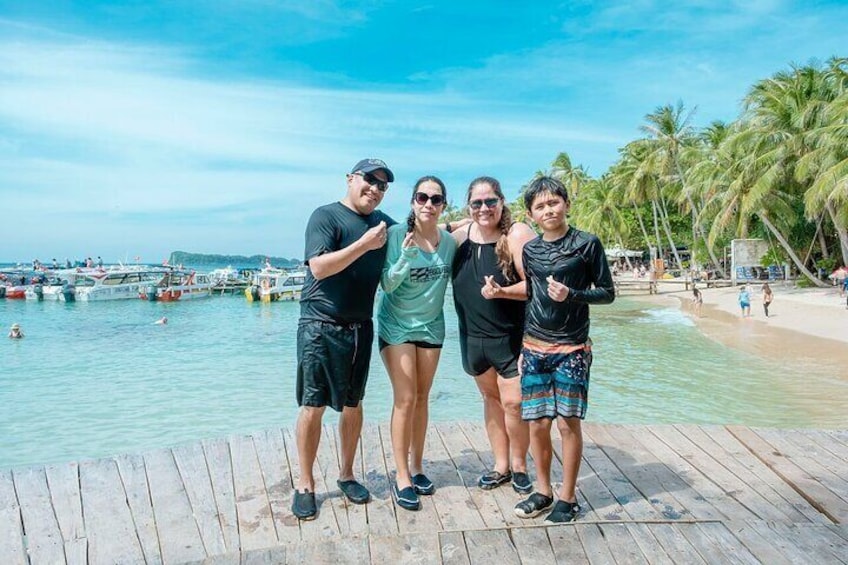 A group of people standing on a wooden dock by a beach with palm trees and boats in the background. 1