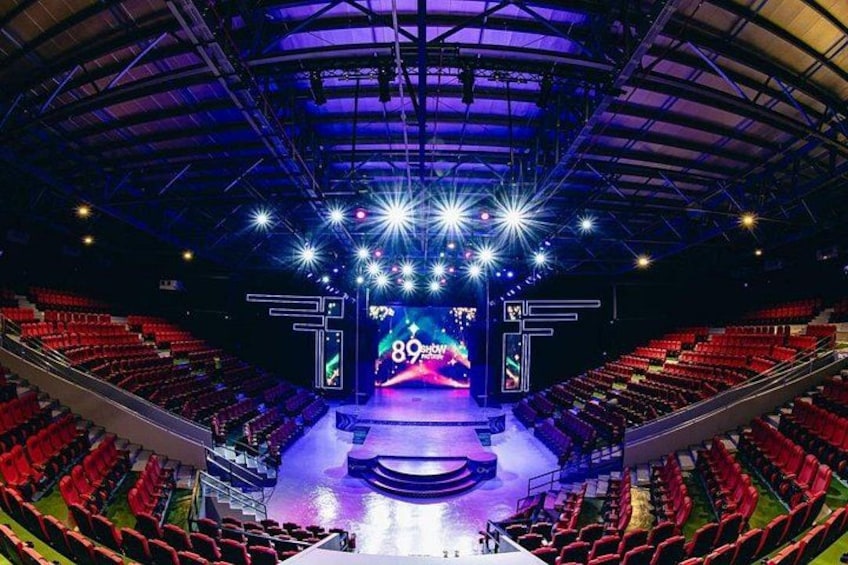 An indoor auditorium with red seating, a stage with a large screen, and bright overhead lights. 4