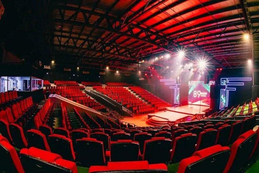 An indoor auditorium with red seats, a stage, and a large screen displaying '89th'. 2