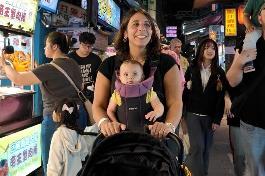 A woman with a baby in a carrier, a crowded street with neon signs, and people walking. 1