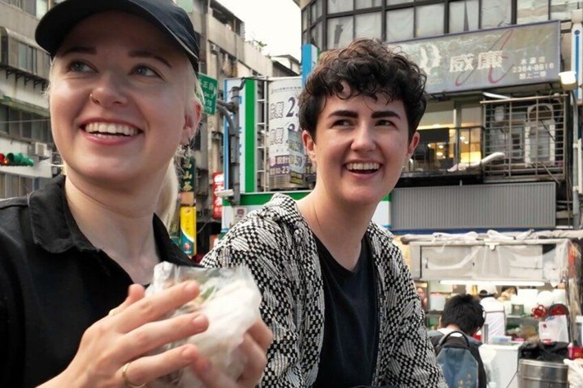 Two people in an urban setting with buildings and signage in the background. 3