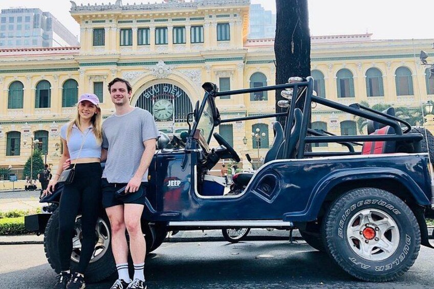 Un fuoristrada blu Jeep con un pneumatico di scorta montato sul retro, parcheggiato di fronte a un edificio con una torre dell'orologio e finestre ad arco. 1