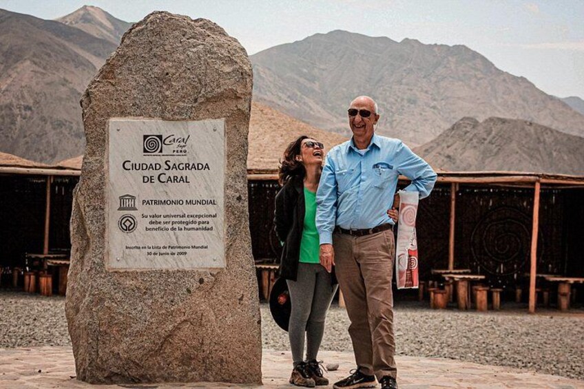 A stone monument with inscriptions, a man and a woman standing beside it, and a mountainous landscape in the background. 1