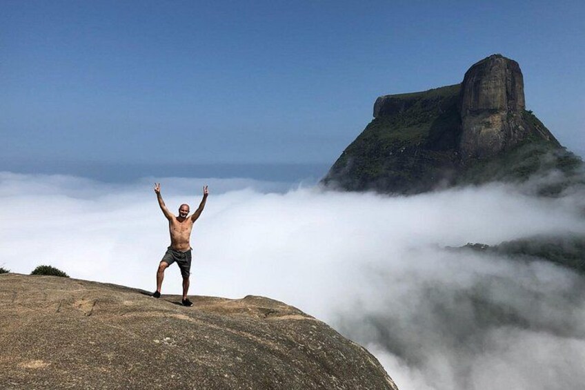 A person standing on a rock with arms raised, a large rock formation in the background, and clouds below. 1