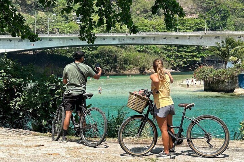 Two people on bicycles by a river with a bridge in the background. 1