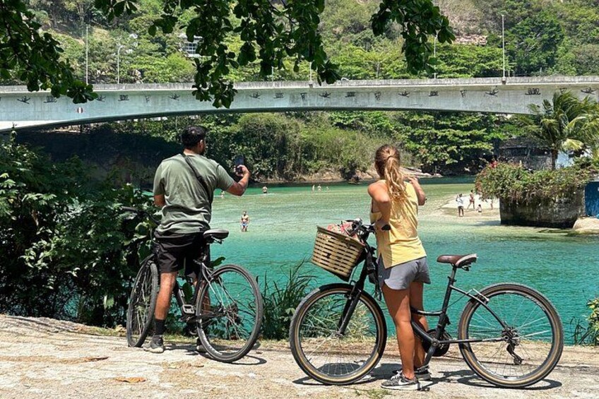 Two people on bicycles by a river with a bridge in the background. 1