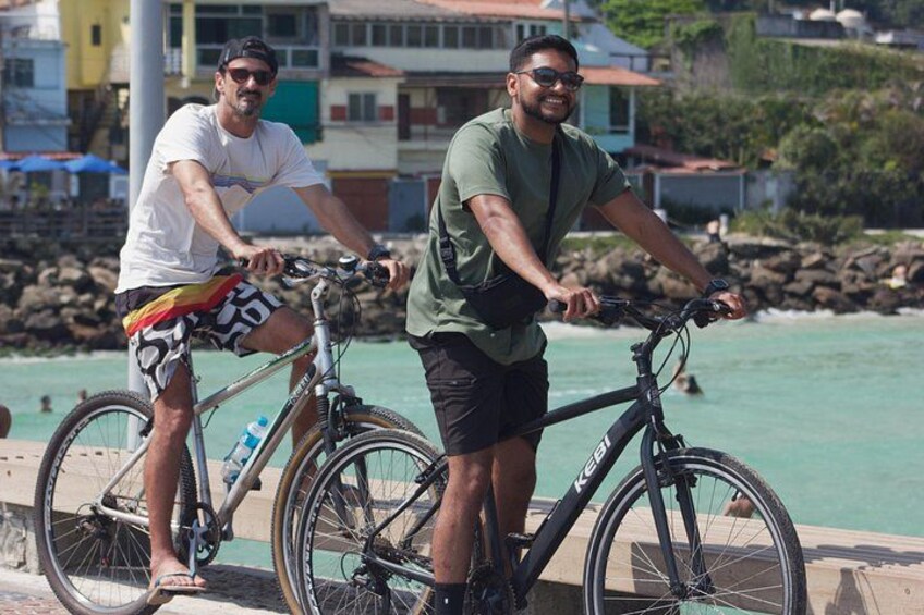 Two people riding bicycles on a waterfront promenade with buildings and a rocky shoreline in the background. 4