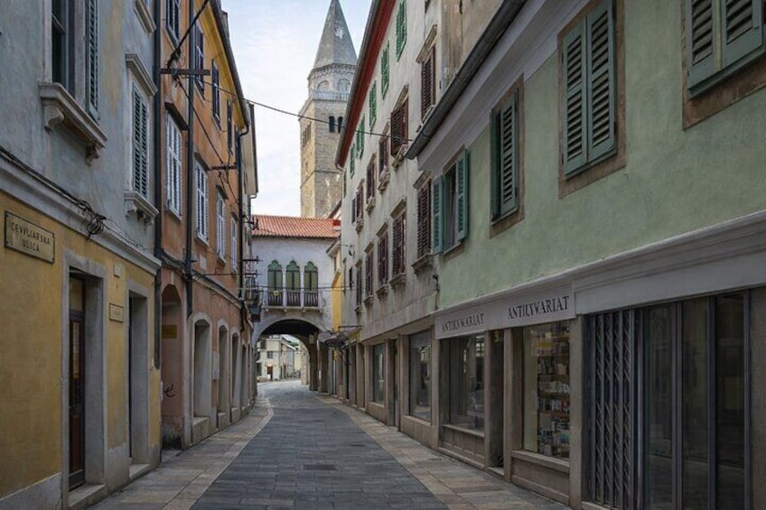 A narrow street with buildings on either side, a tower in the background, and a sign for 'ANTICIVARIAT'. 2