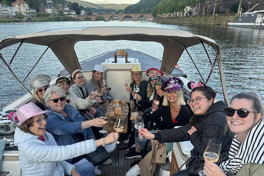 A group of people on a boat, toasting with drinks, with a river and buildings in the background. 5