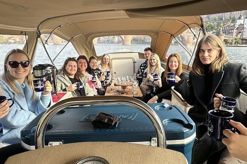 A group of people on a boat with a table in the center, holding mugs. 3