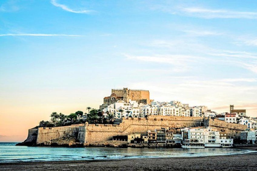 A coastal cityscape with a prominent castle on a hill, a beach in the foreground, and a clear sky. 1