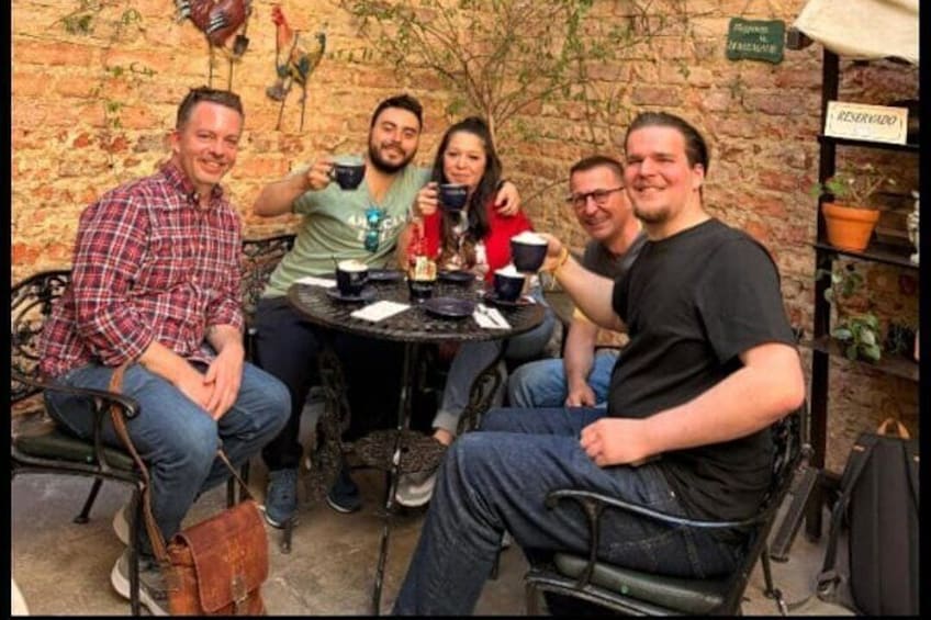 Four people sitting at a round table outdoors with a brick wall and plants in the background. 3