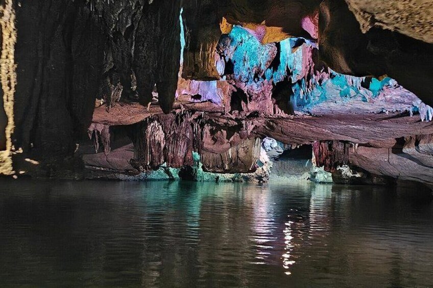 Cueva con estalactitas y estalagmitas de colores, un cuerpo de agua que refleja el interior de la cueva y una estructura de madera. 5