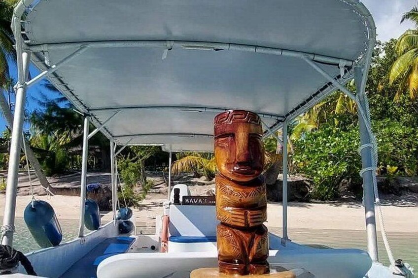 A boat with a wooden tiki head sculpture on the bow, a white canopy, and a beach with palm trees in the background. 2