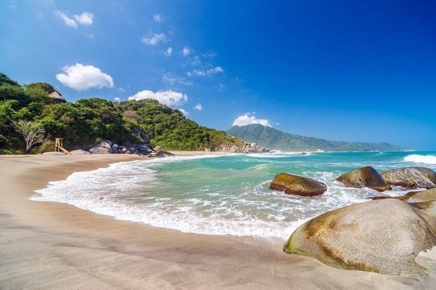 A beach with clear turquoise water, large rocks, and a forested hillside. 4