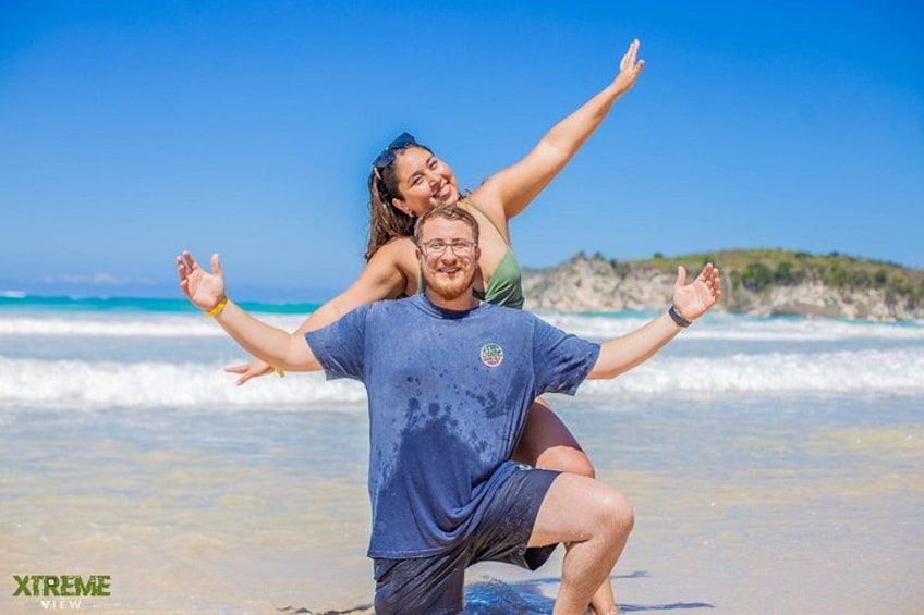 A man carrying a woman on his back on a beach with clear skies and ocean waves. 5