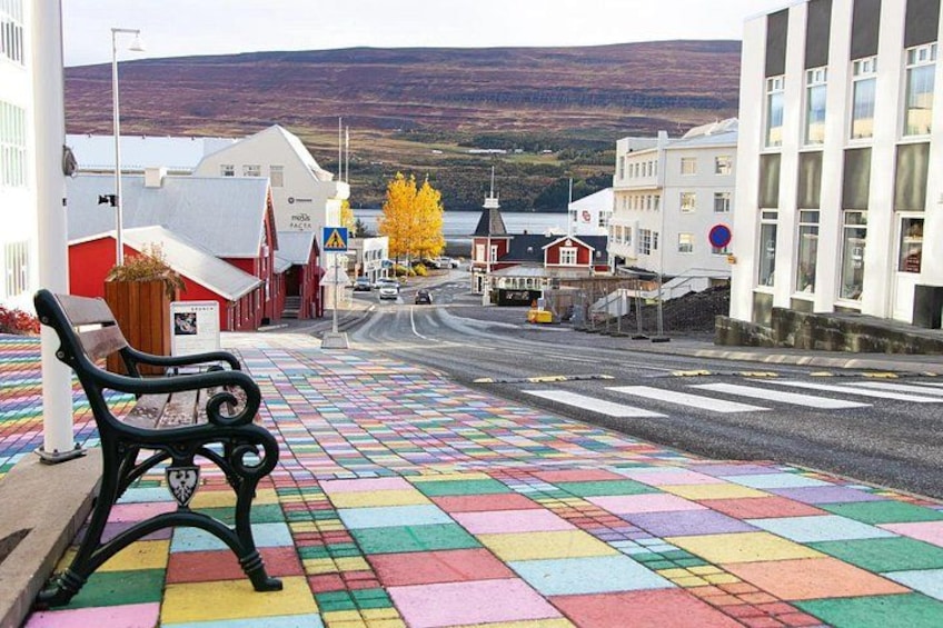 A colorful tiled pedestrian walkway, a bench, and buildings with a view of a hill and water in the background. 3