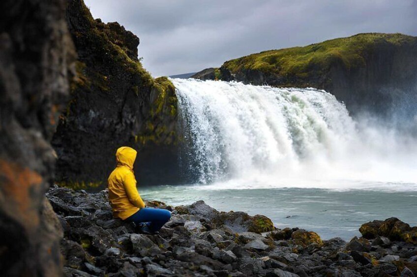 A person in a yellow jacket sitting on rocks by a waterfall. 3