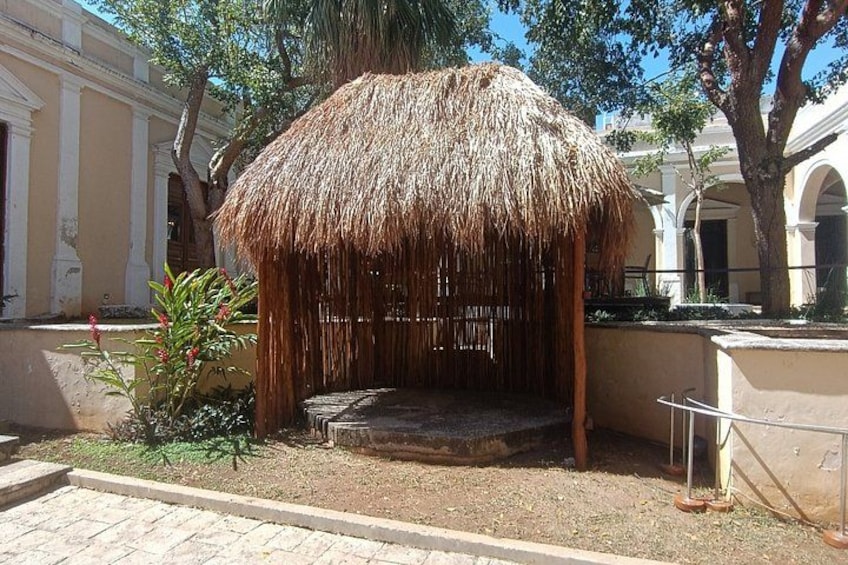 A thatched-roof structure with steps leading up to it, surrounded by trees and a building with columns. 1