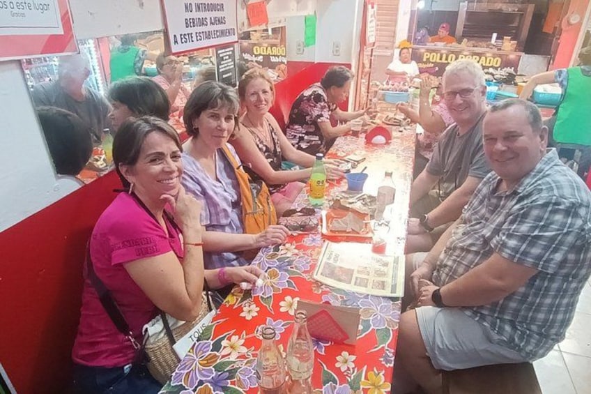 A group of people seated around a table with various items on it, in a brightly lit indoor setting. 4