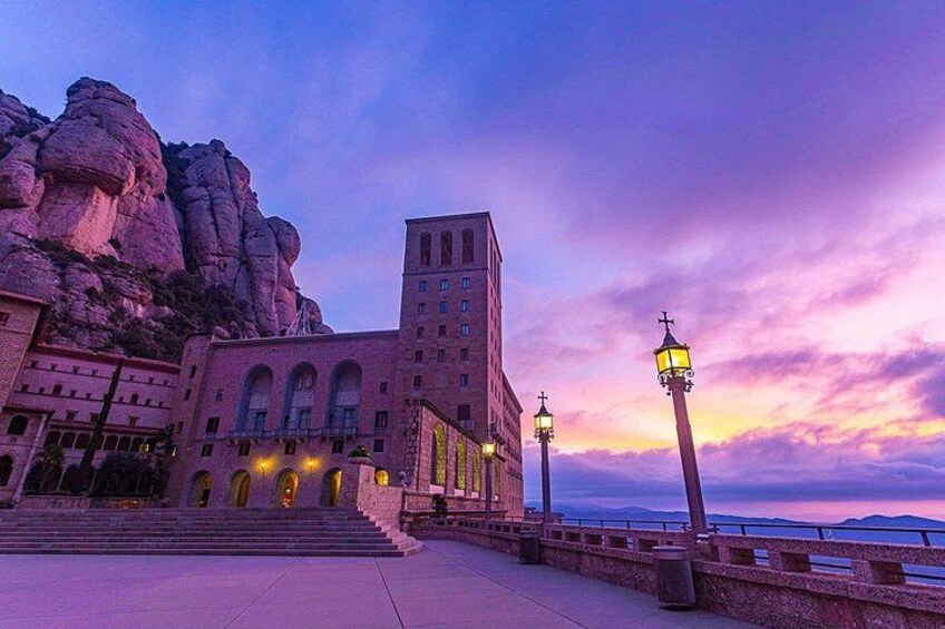 A building with arched windows and a tower, a rocky cliff, and a sunset sky. 2
