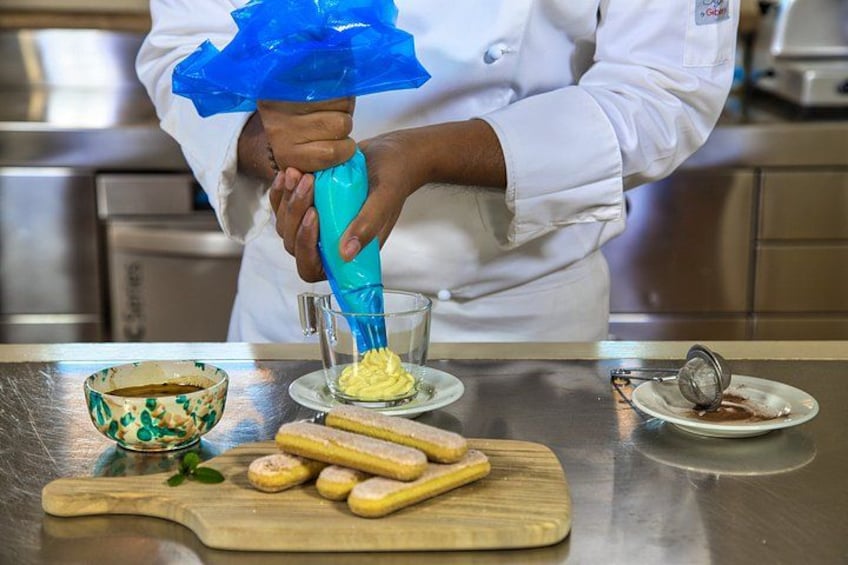 A chef in a white coat using a blue piping bag to fill a glass with a yellow substance, possibly butter, on a stainless steel countertop with a wooden cutting board and pastry dough. 4