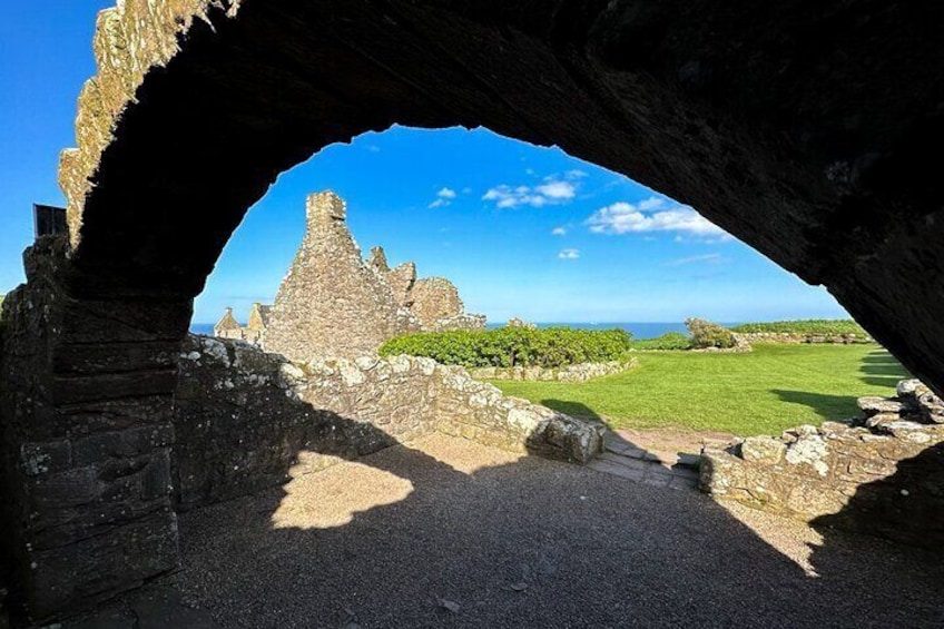 An ancient stone structure with a large archway, a grassy field, and a distant view of the ocean. 2