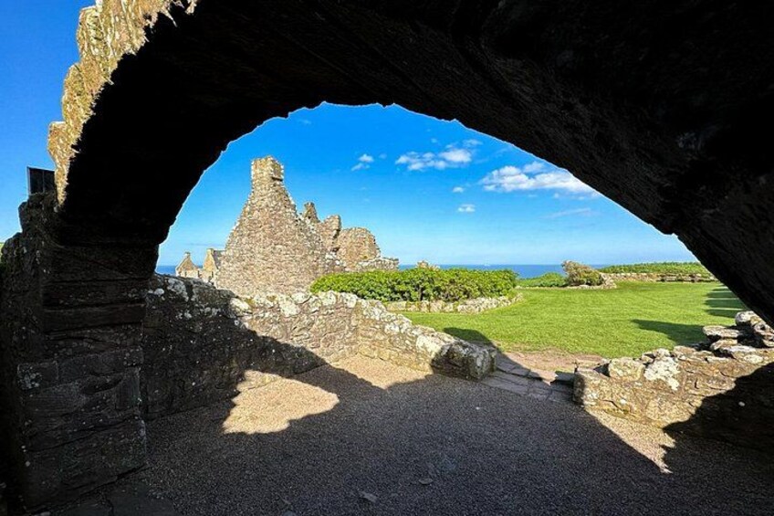 An ancient stone structure with a large archway, a grassy field, and ruins in the background. 2