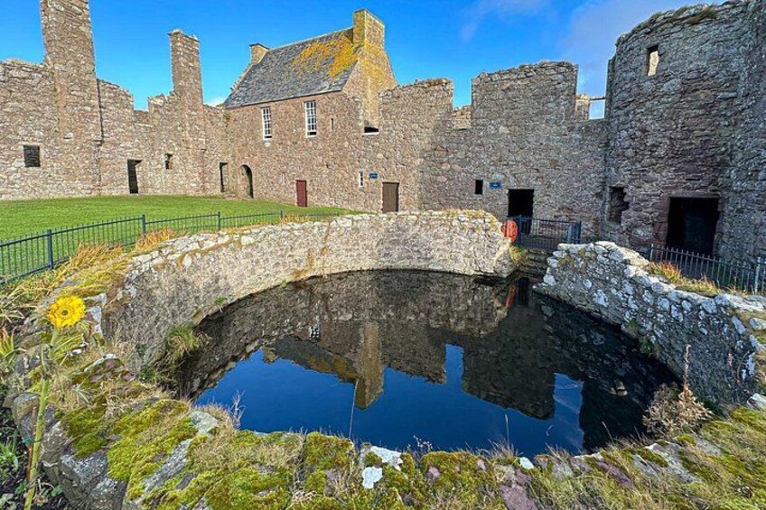 A stone bridge over a reflective water body, surrounded by historic stone buildings with a grassy area in the foreground. 3