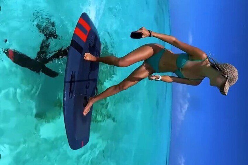 A person in a bikini and hat holding a surfboard, standing in clear turquoise water. 2