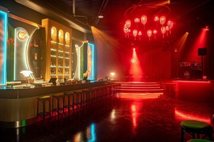A bar with a counter, stools, and a stage with red lighting and hanging lanterns. 1