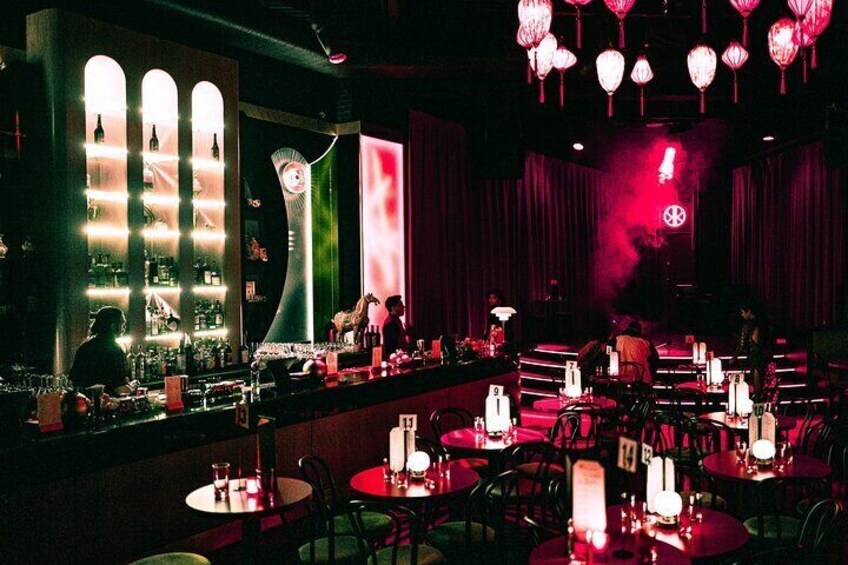A dimly lit bar with tables, hanging lanterns, and a stage with a red light. 5