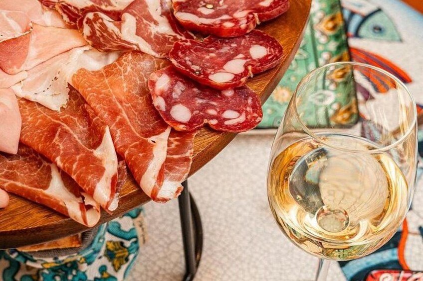 A wooden board with various types of cured meats, a glass of white wine, and a patterned tablecloth. 2