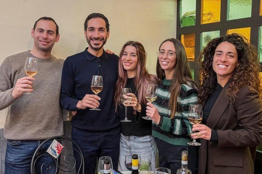 A group of people holding wine glasses, standing indoors with a table of wine bottles and glasses in front of them. 1