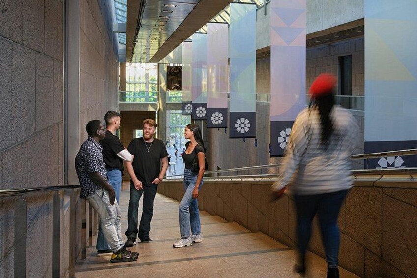 A group of people standing in a corridor with a glass ceiling, a patterned wall, and a person walking by. 4