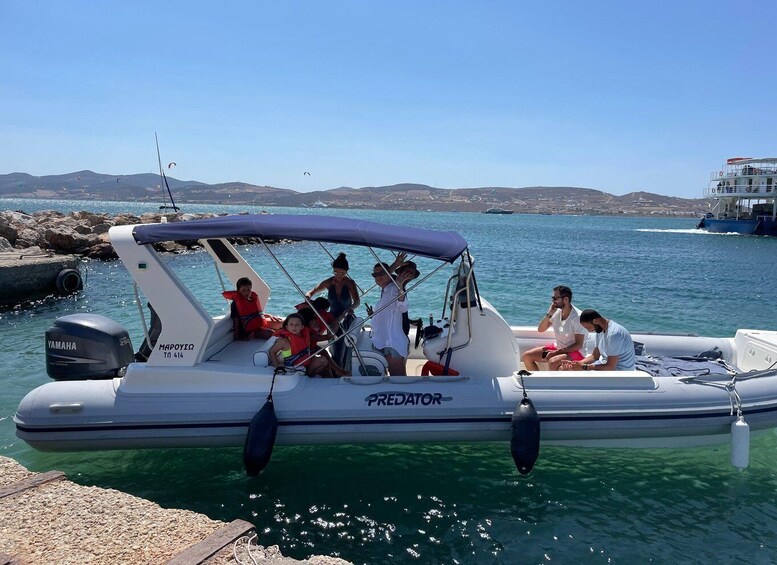 A small inflatable boat with people on board, anchored near a rocky shore with clear water and distant hills. 1