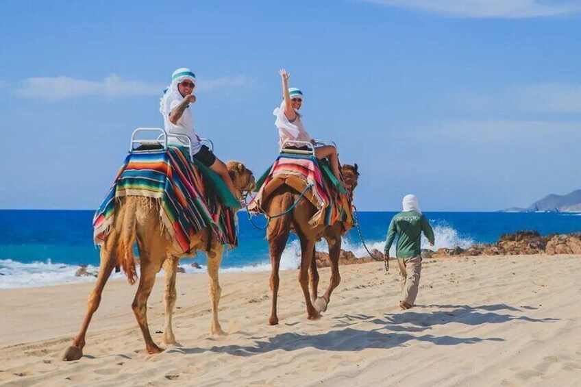 Two camels with riders on a beach, a person walking behind them, and the ocean in the background. 1