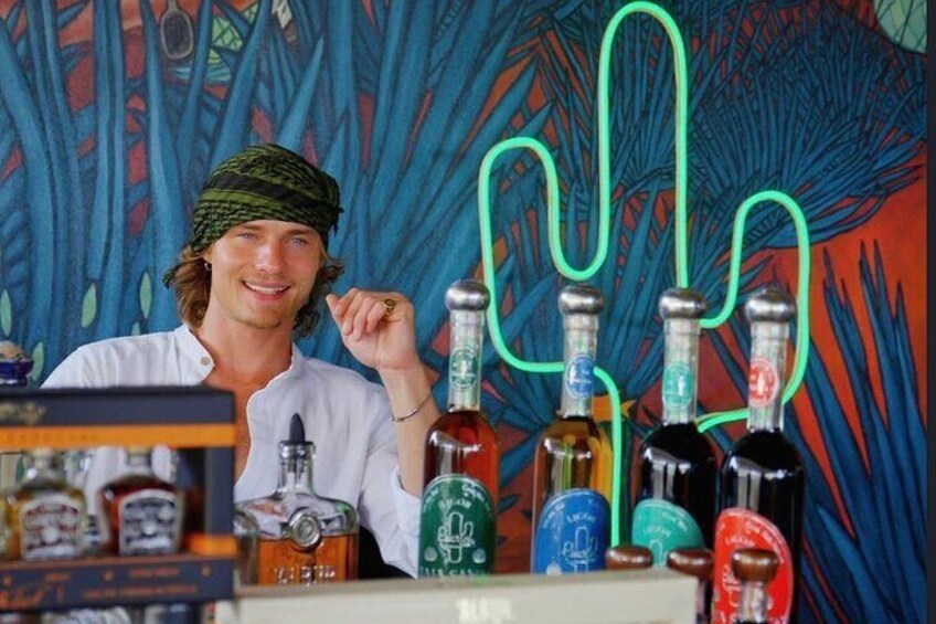 A person in a white shirt with a green headscarf, standing in front of a colorful mural. There are several bottles of alcohol on the counter in front of the person. 2