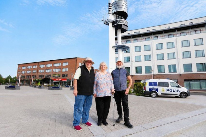 Three people standing in a public square with a modern building and a police van in the background. 3