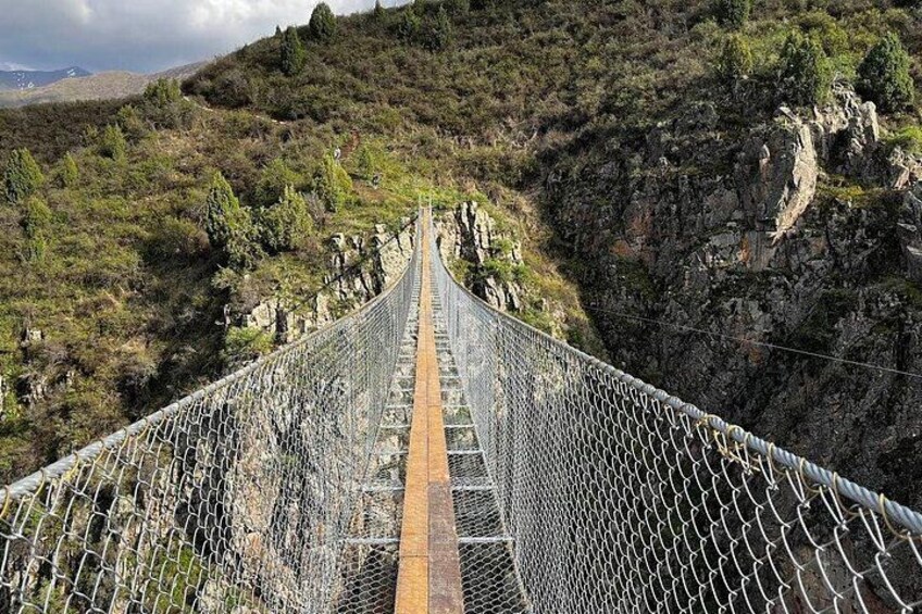 A suspension bridge with a chain-link fence, surrounded by rocky terrain and sparse vegetation. 3