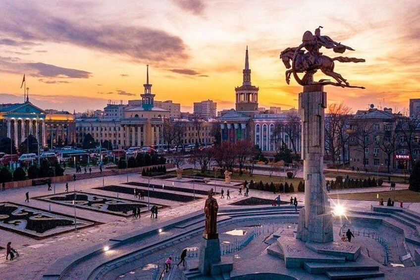 A city square with a central monument, surrounding buildings, and a sunset sky. 3