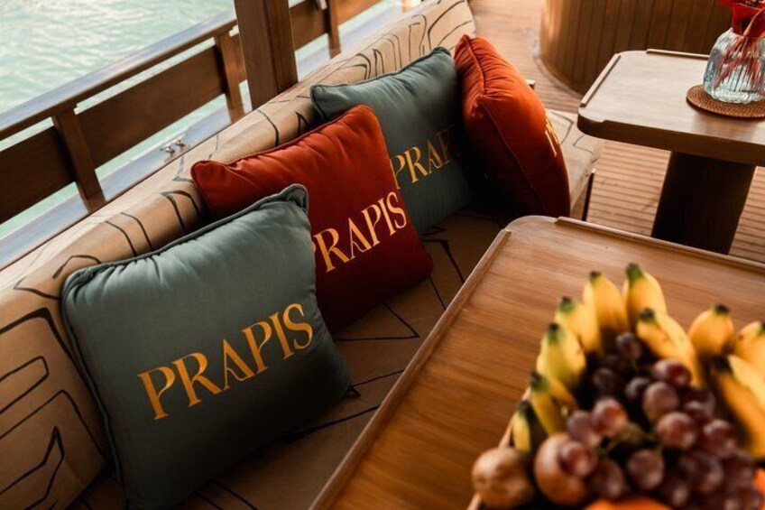 Une terrasse dotée de sièges rembourrés, de coussins ornés du mot « PRAPIS » et d’une table sur laquelle sont disposés des fruits. 2