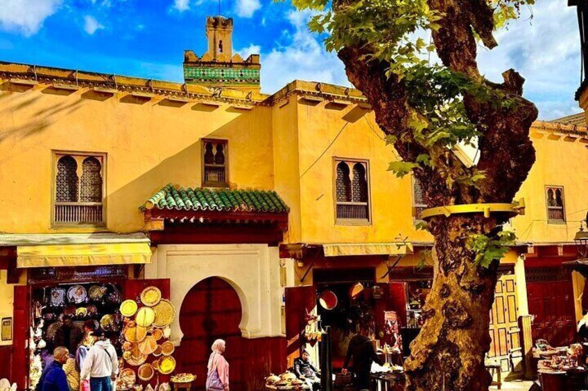 A bustling street market with yellow buildings, a large tree, and people shopping. 1