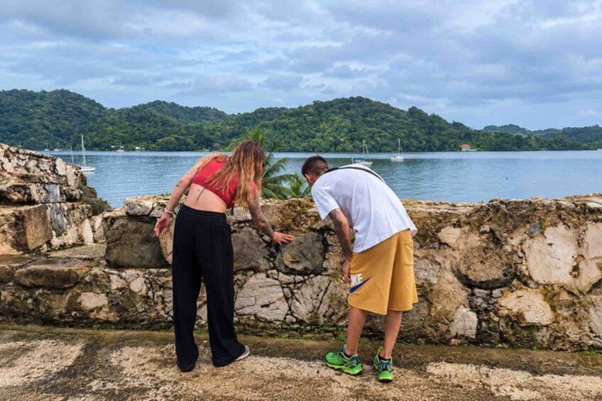 Two people standing by a stone wall overlooking a body of water with boats and a forested island in the background. 5
