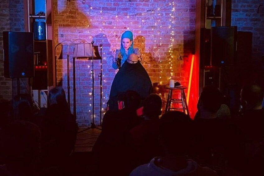 A person performing on stage with a microphone stand, audience in front, and brick wall with string lights. 2