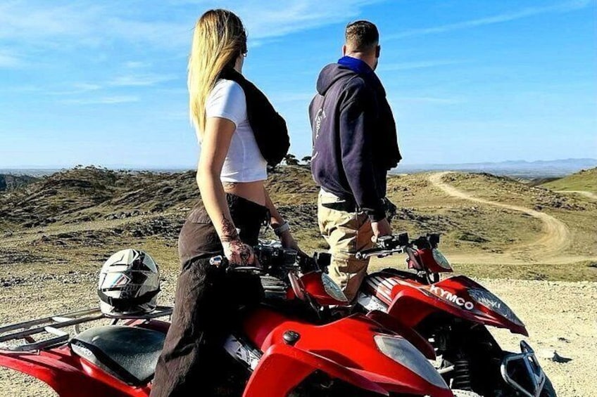 Two people standing on a red ATV in a hilly, open landscape. 4