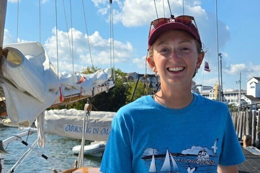 A person on a boat with a sail, docked at a marina with other boats and houses in the background. 5