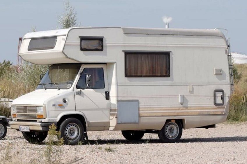 Una caravana blanca estacionada sobre grava, con capota, ventanas laterales y una pequeña ventana trasera. 2